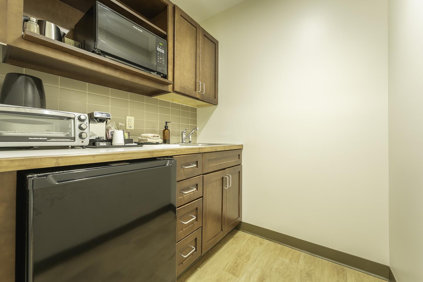 Small kitchenette with wooden cabinets, microwave, fridge, and countertop appliances.