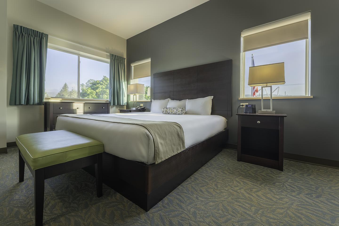 Bright hotel room with a large bed, green bench, and two windows with curtains.