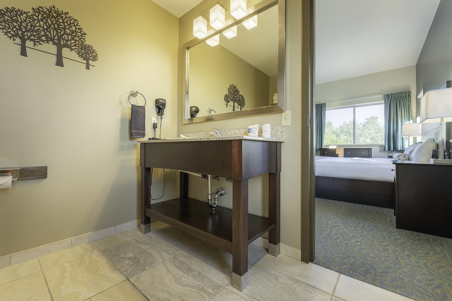 Hotel room bathroom with modern vanity, large mirror, and view into bedroom.