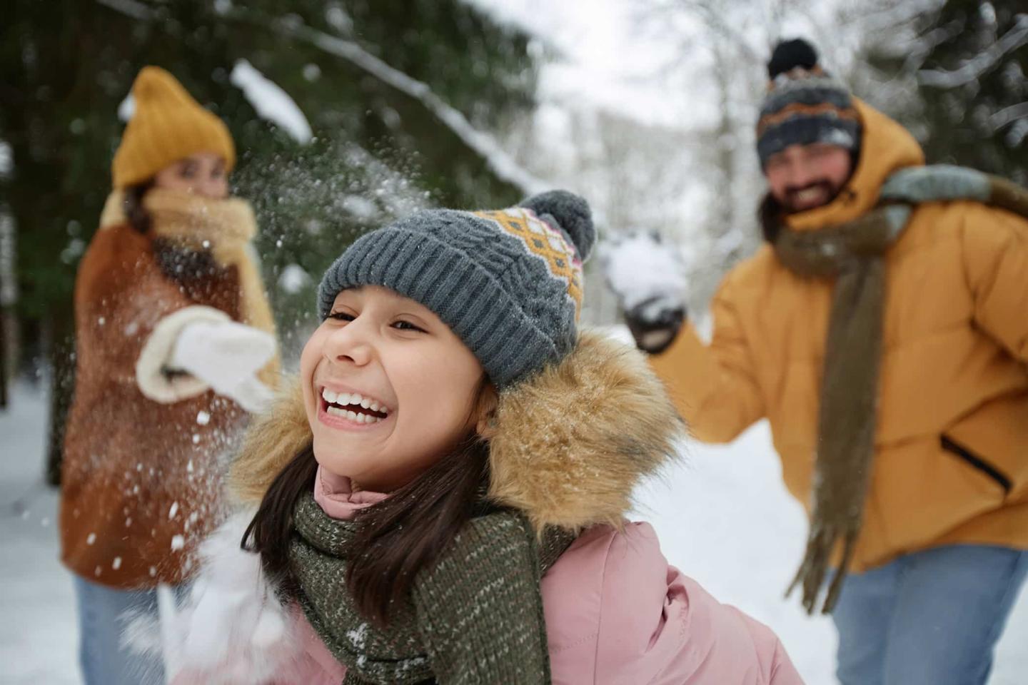 Child laughing during snowball fight with adults in winter clothing.