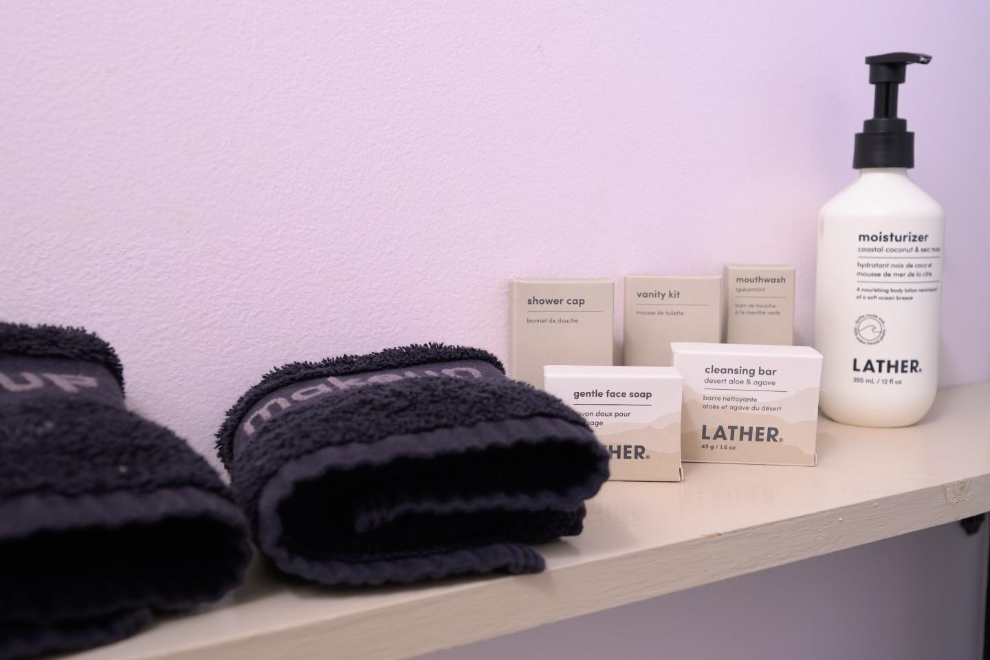 Black towels and LATHER toiletries on a shelf against a pale wall.