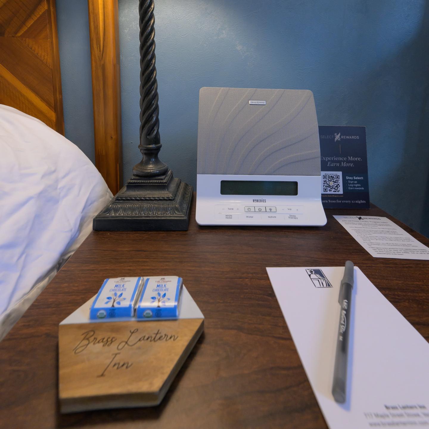 Hotel nightstand with clock, notepad, pen, and chocolates.