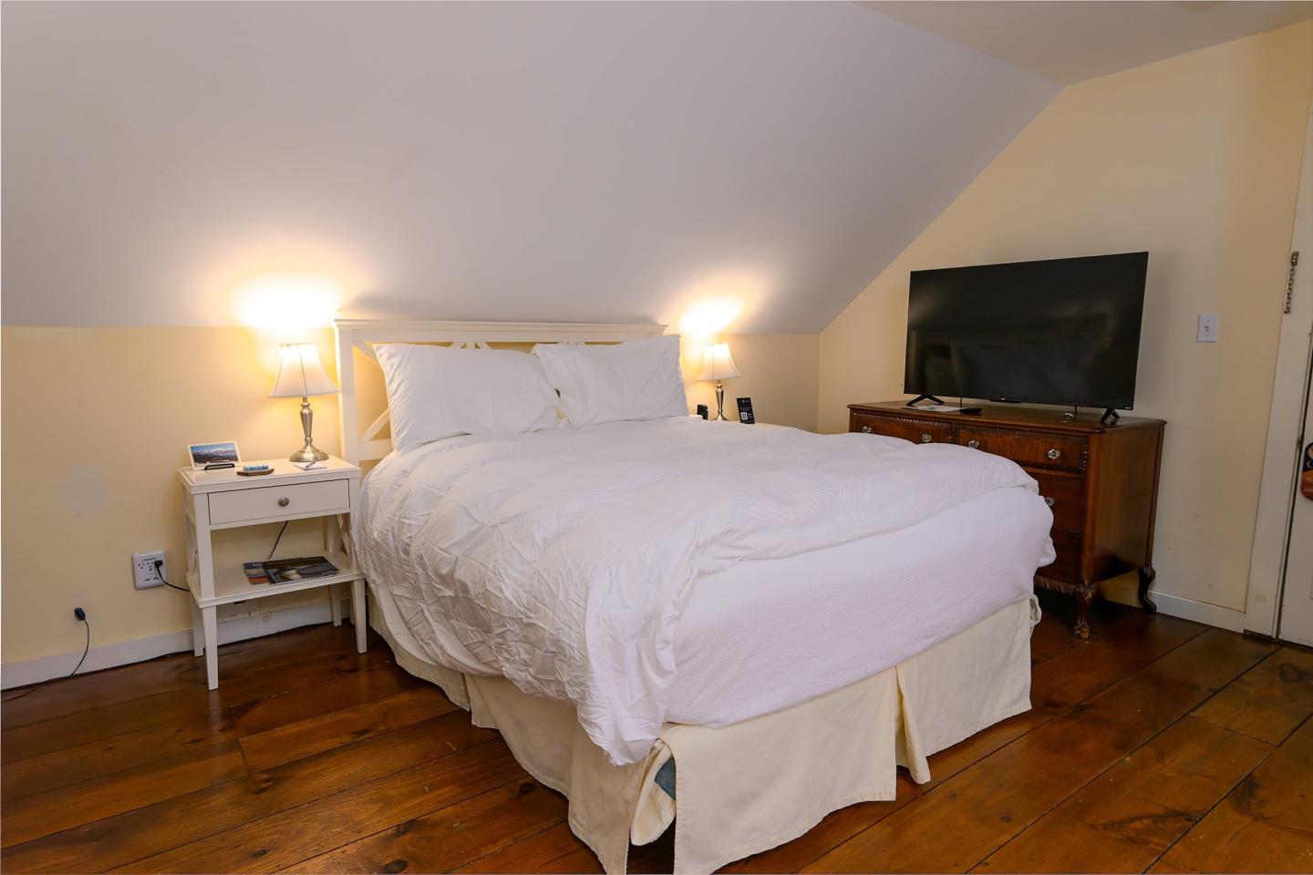 Cozy bedroom with white bed, wooden floor, and TV on dresser. Warm lighting from lamps.