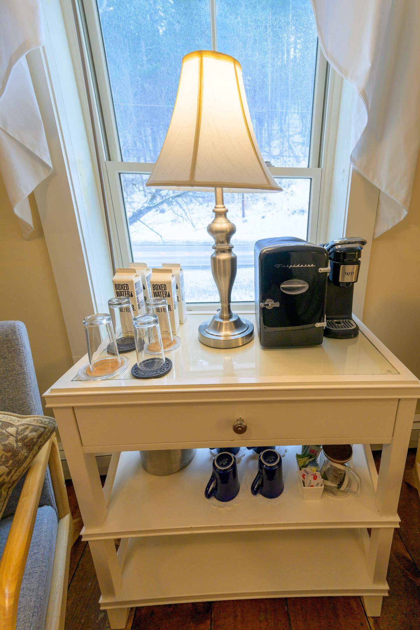 Table with a lamp, coffee maker, glasses, and mugs by a window.