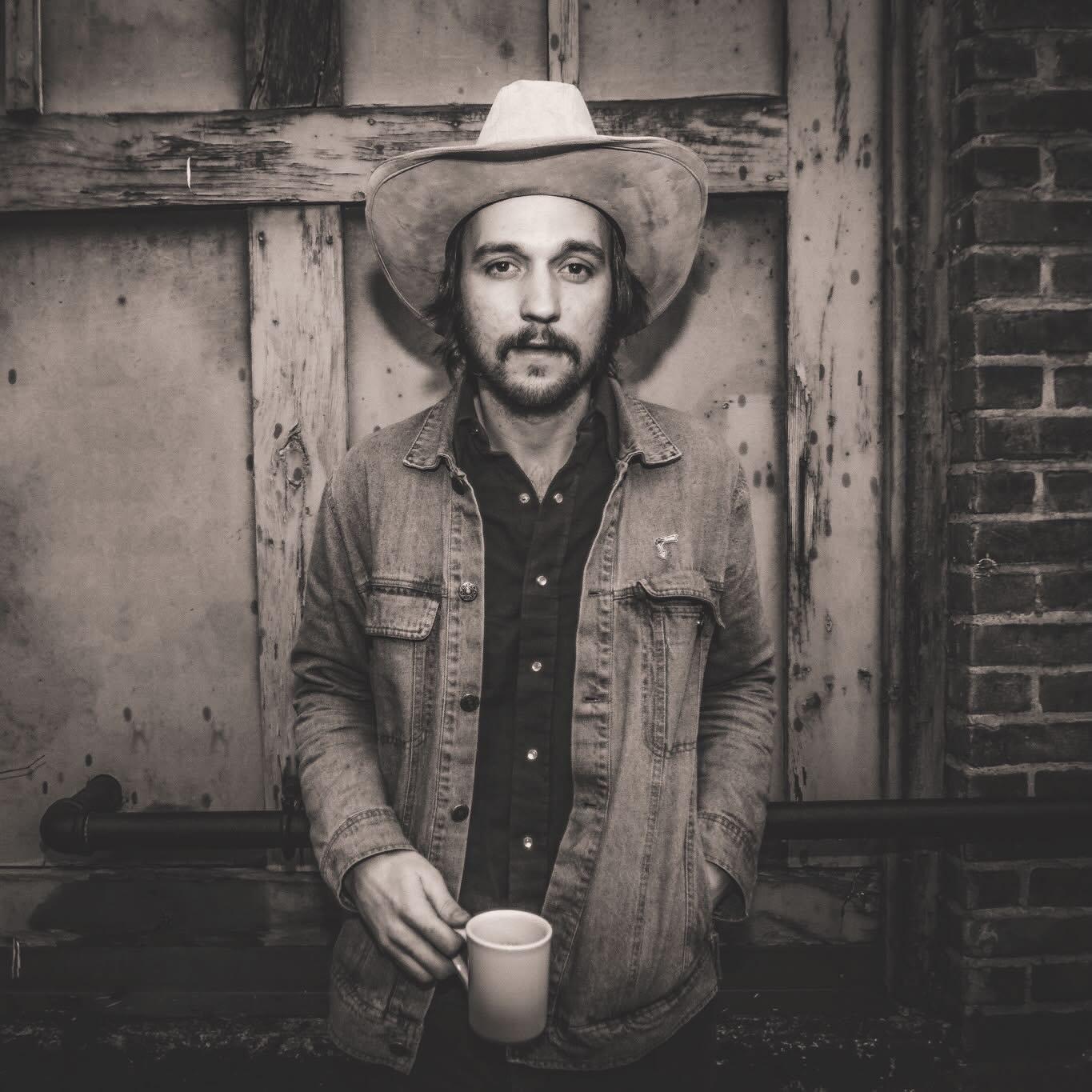 Man in a cowboy hat holding a mug, wearing a denim jacket, black and white photo.