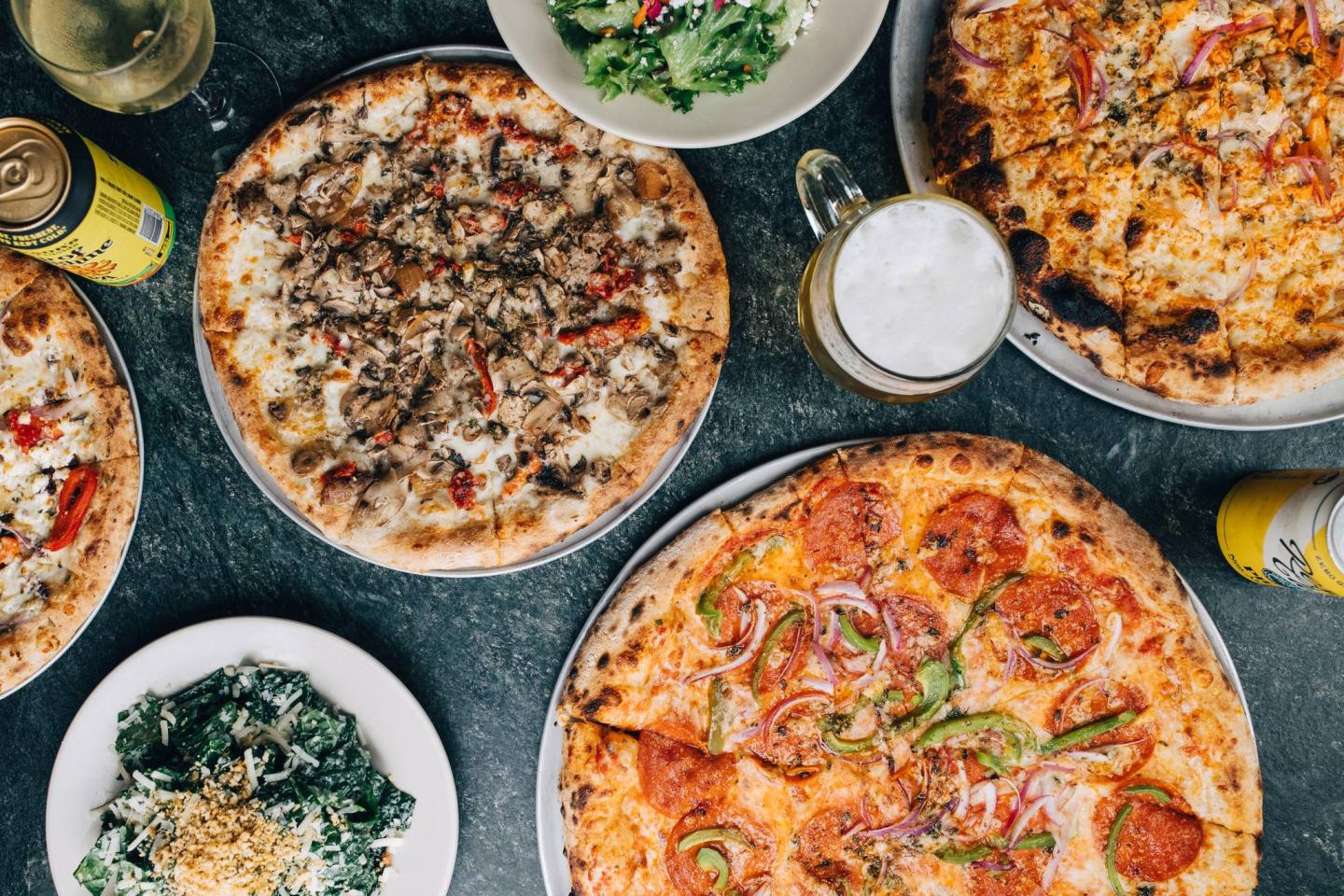Pizzas with various toppings, salads, and drinks on a dark table.