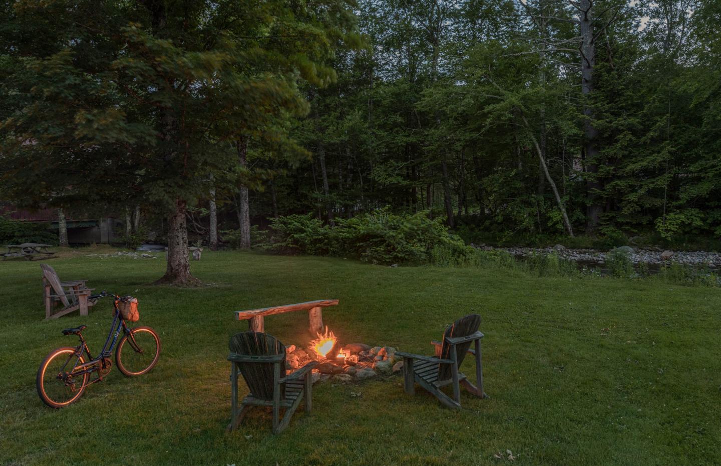Campfire with chairs on a grassy area, bicycle nearby, surrounded by trees.