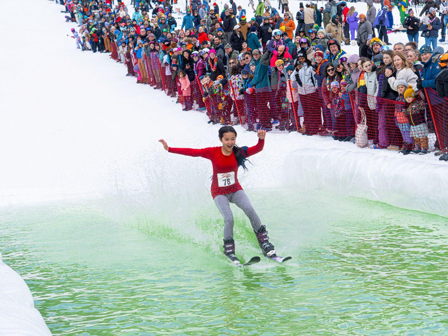 Skier in red crossing a green water pit, surrounded by a cheering crowd.