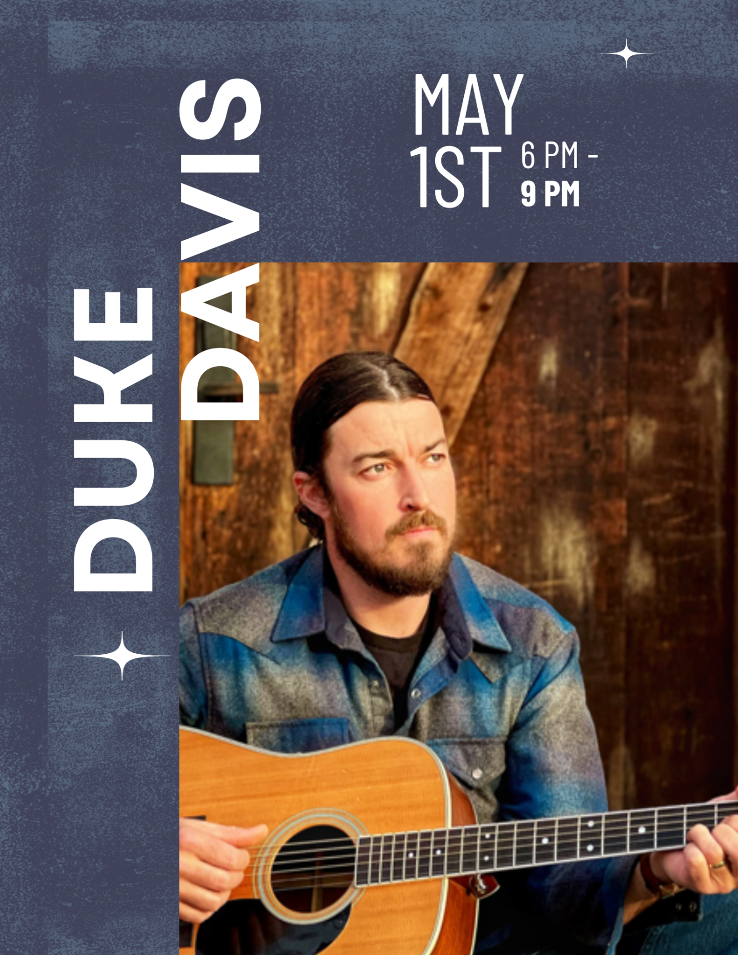 Musician with guitar, wearing a blue plaid shirt, seated in a rustic setting.