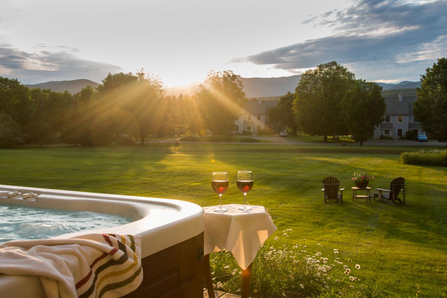 Hot tub in a garden at sunset, with wine glasses on a table, facing Adirondack chairs.