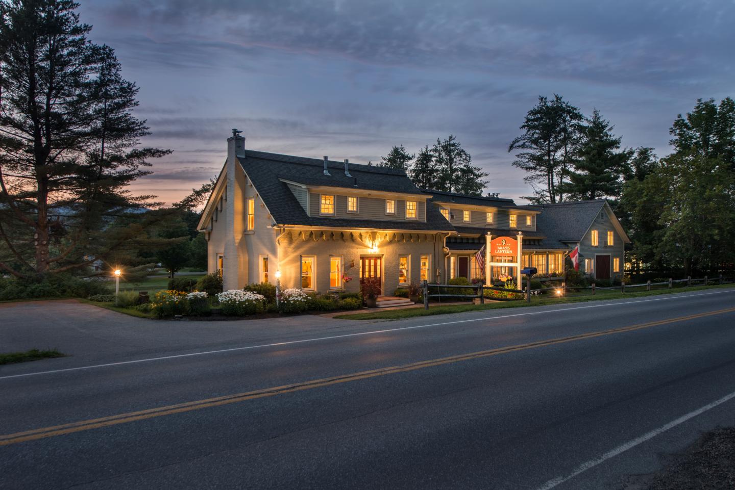 Cozy house with warm lights at dusk, trees in background, road in foreground.