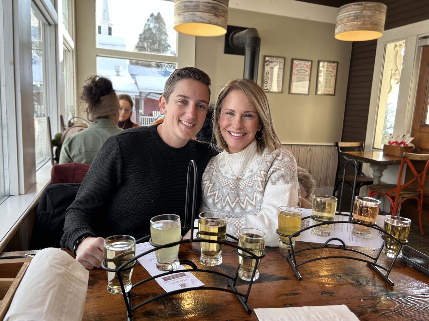 Two people smiling at a table with glasses of hard cider inside cold hollow cider mill.
