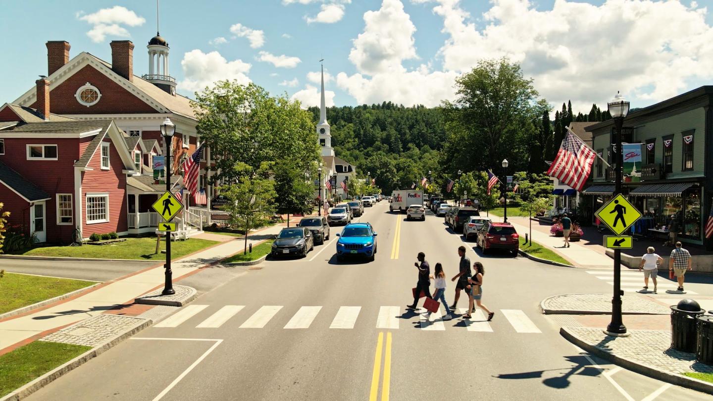 Small town street with pedestrians crossing, flanked by brick buildings and greenery.