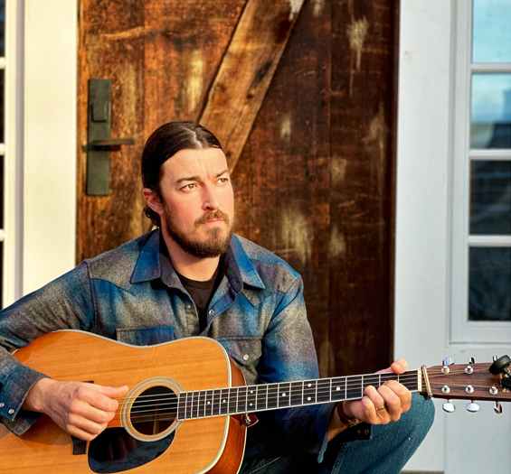 Man with a beard playing a guitar, seated by a wooden door.