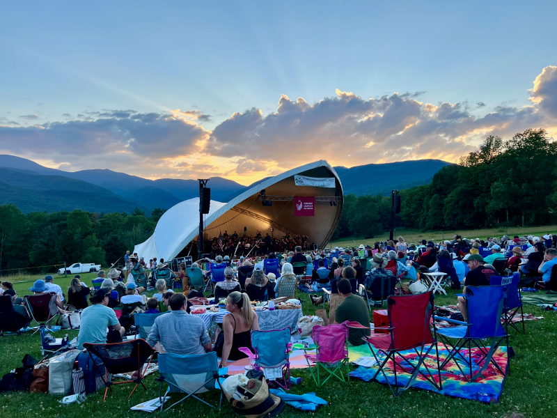 Outdoor concert at sunset with an audience, mountains, and a cloud-filled sky.