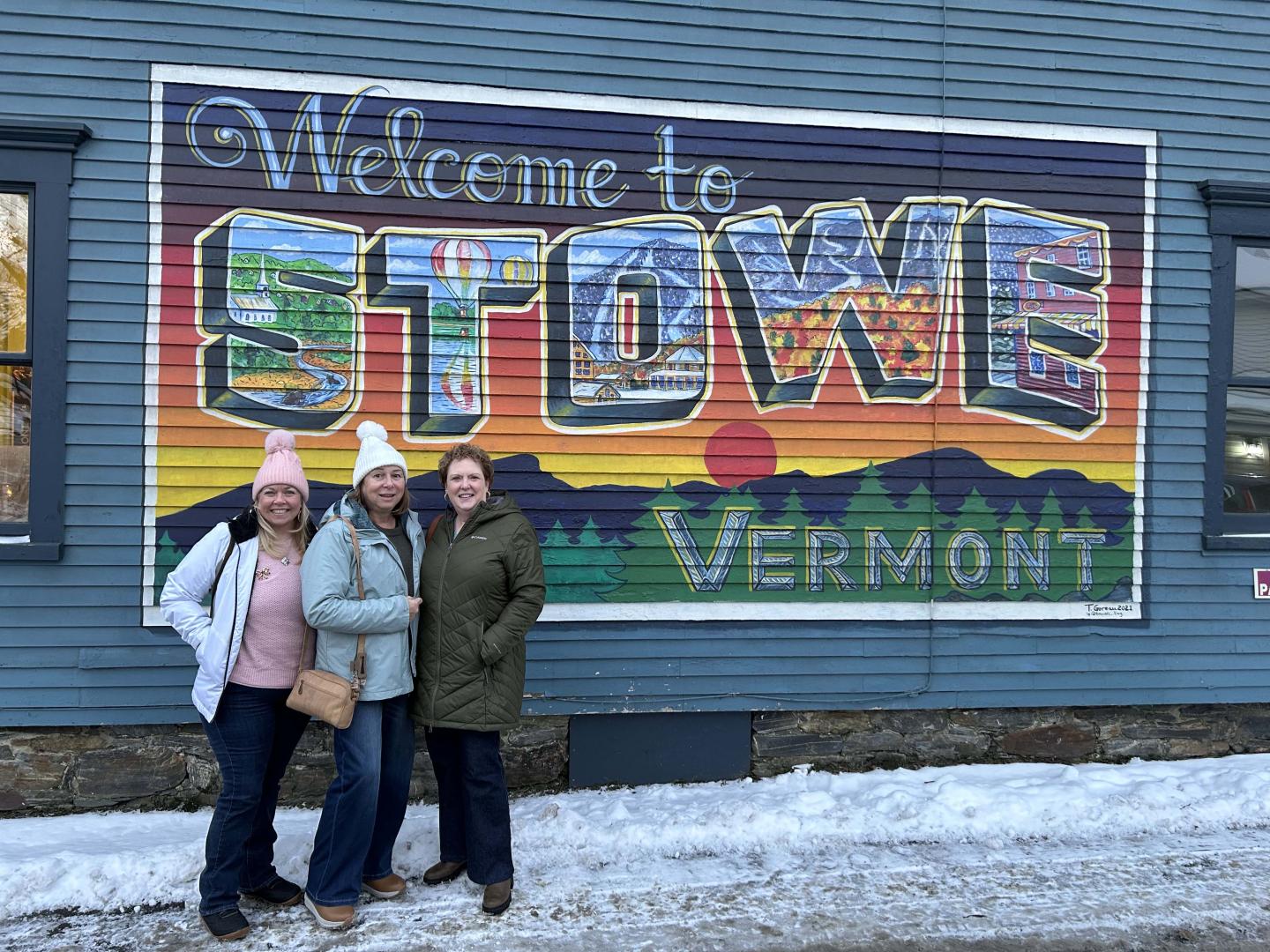 Three people smiling in front of a colorful mural reading "Welcome to Stowe Vermont."
