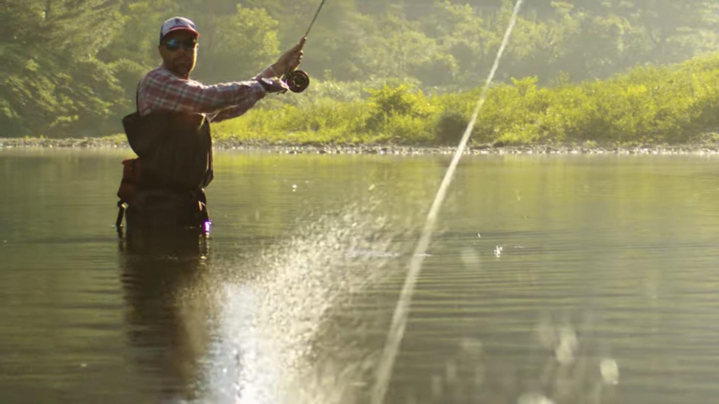 Man fly fishing in a river, surrounded by trees and sunlight.