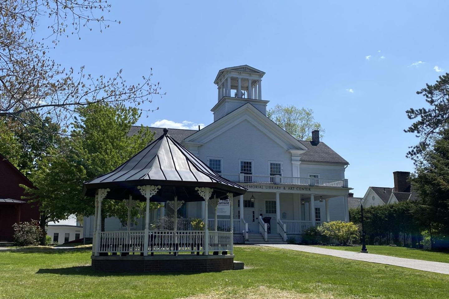 Gazebo on grassy lawn with large white house in background under blue sky.