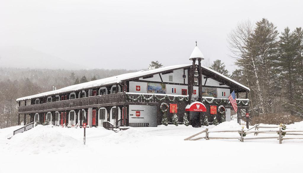 Snow-covered lodge with wooden fence, surrounded by trees, overcast sky.