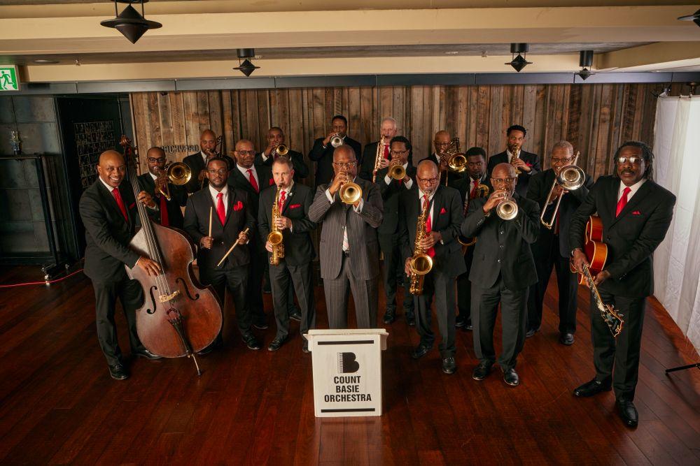 Orchestra in suits with brass instruments, posed on a wooden stage.