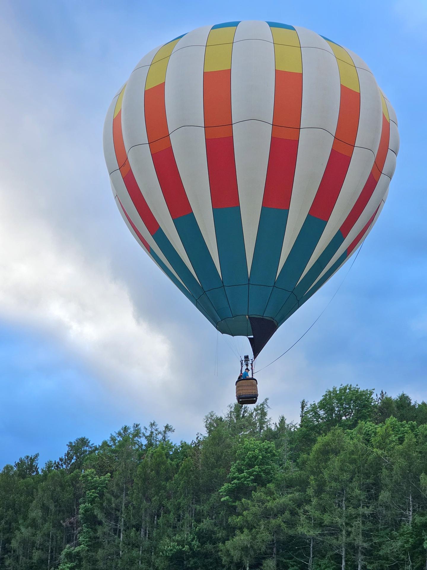 Colorful hot air balloon above green treetops with a cloudy sky.