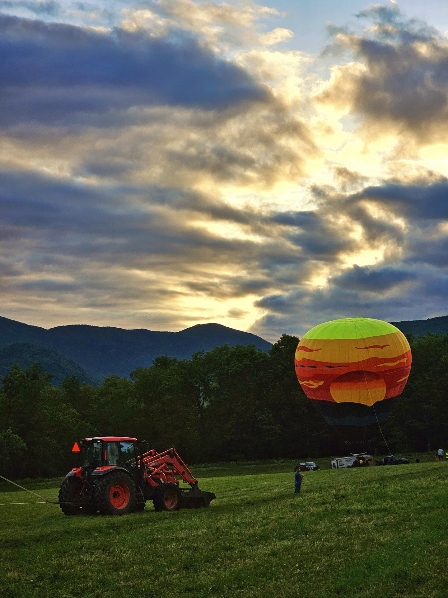 Tractor near colorful hot air balloon at sunrise, mountains in background.