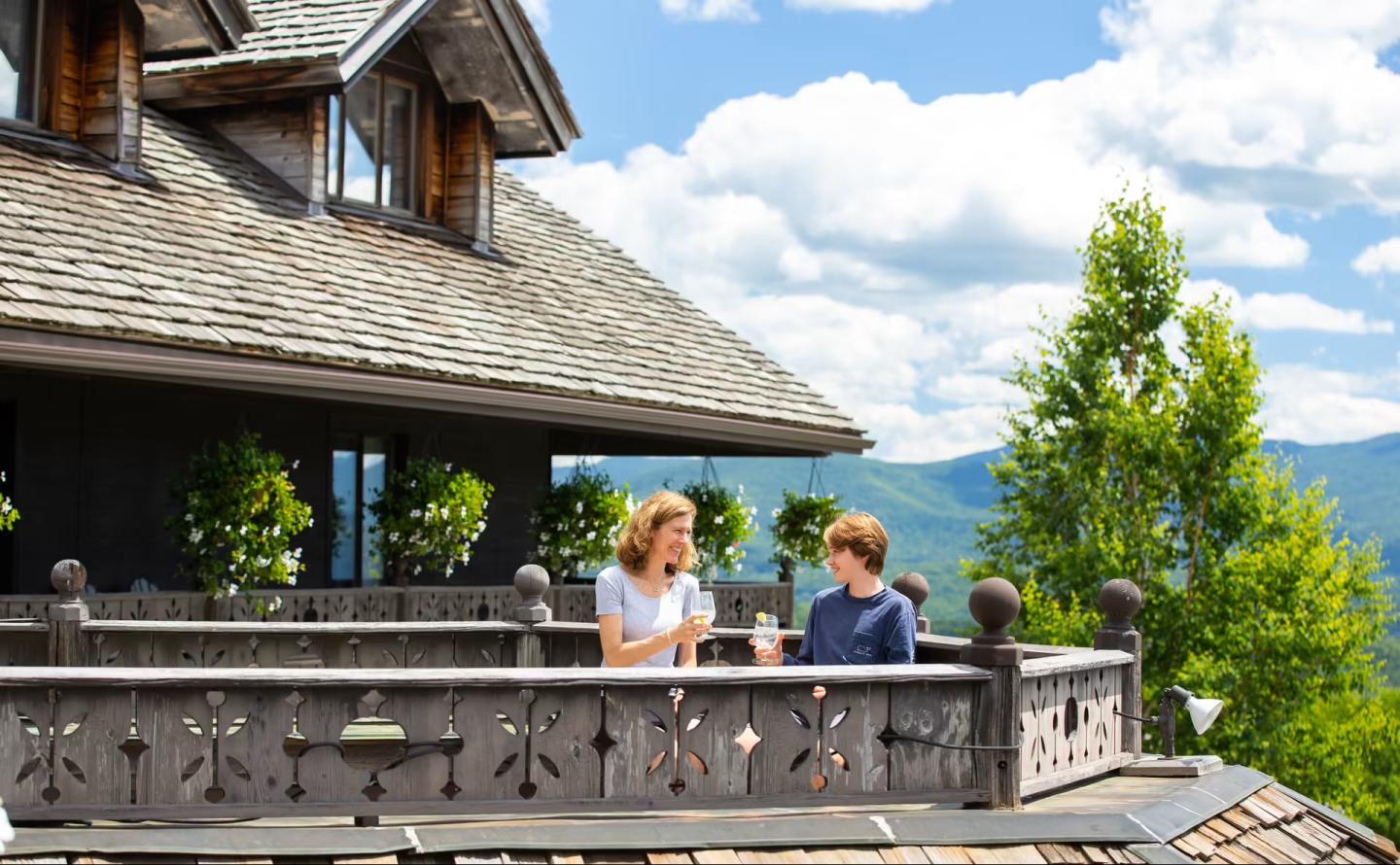 Two people chat on a wooden balcony, surrounded by mountains and trees.