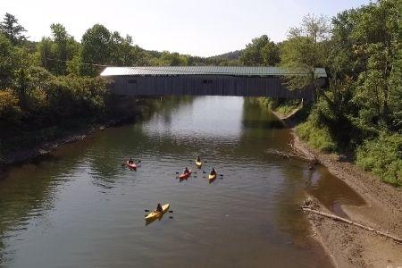 Kayakers paddle under a wooden covered bridge on a calm river surrounded by trees.