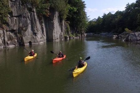 Kayakers on calm river surrounded by rocky cliffs and lush trees.