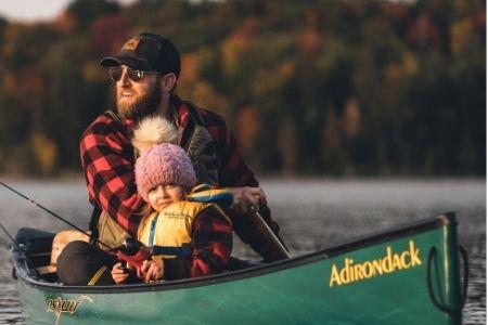 Father and child in a canoe on a calm lake, wearing warm clothing, surrounded by autumn trees.