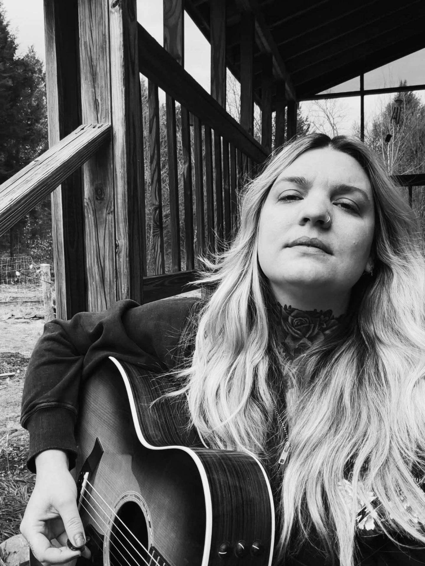 Black and white photo of a woman with a guitar, seated outdoors near a wooden structure.