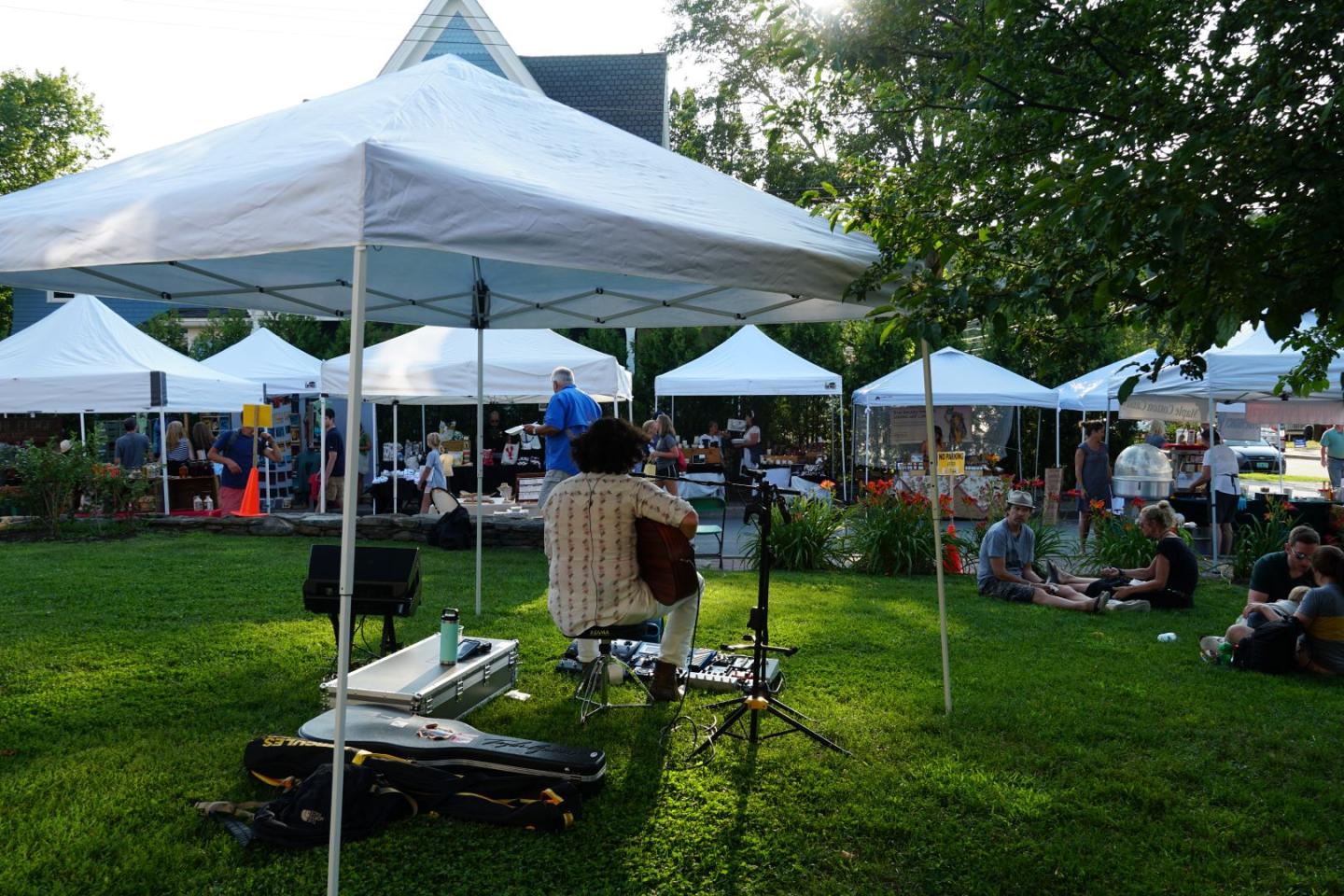 Outdoor market with musician playing under a white tent, surrounded by people.