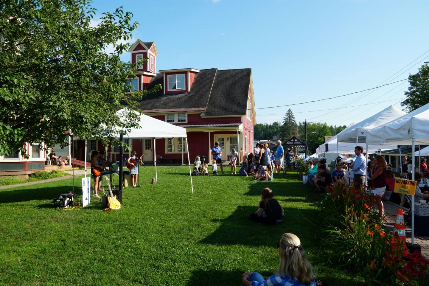Festival on a sunny day, with people sitting on grass near red house and white tents.