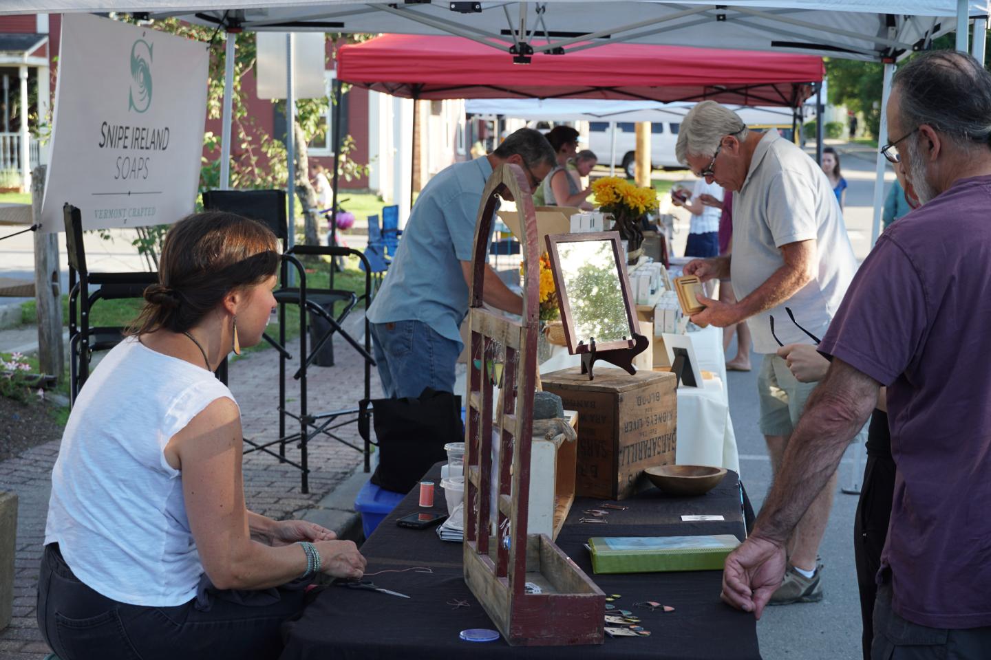 Outdoor market with people browsing crafts under a red tent.