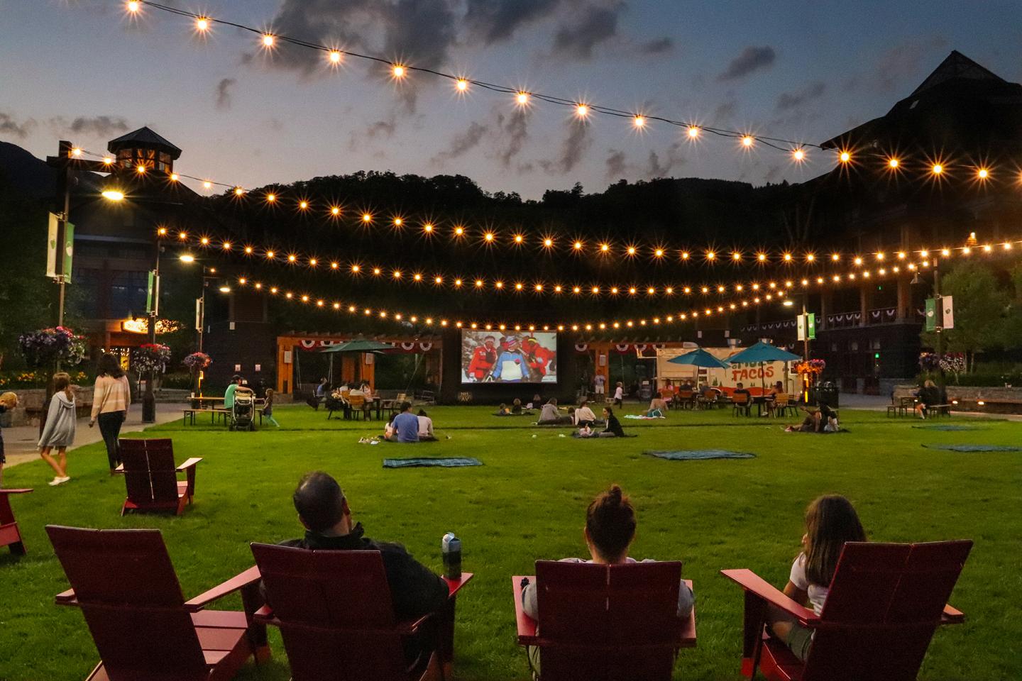 Outdoor movie night with string lights at dusk, people in chairs on grass.