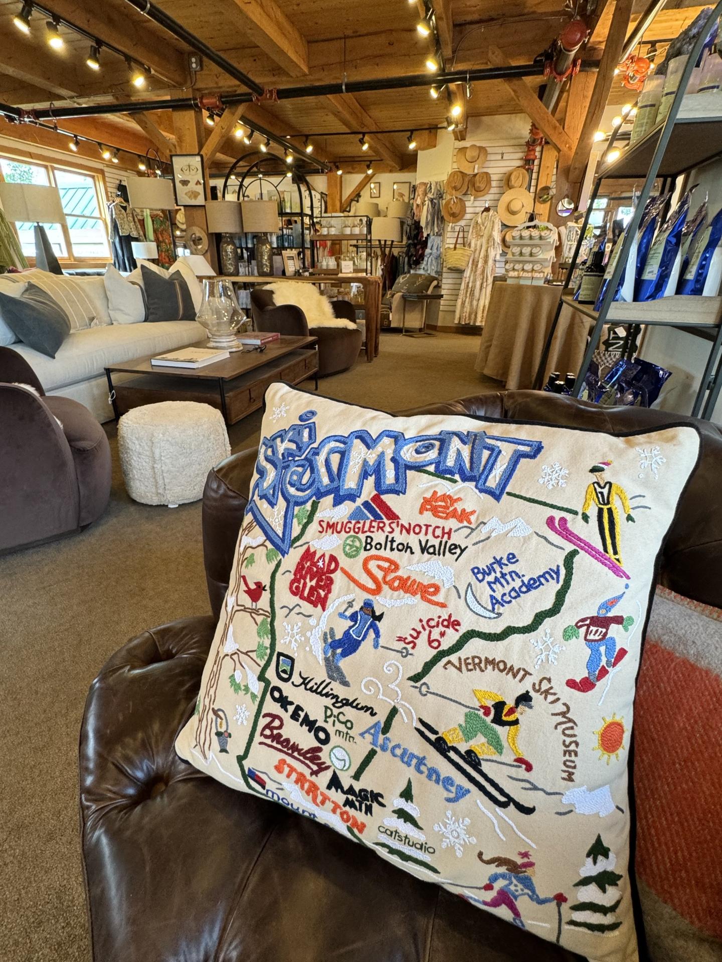 Decorative Vermont map pillow on a leather chair in a cozy, wooden store setting.