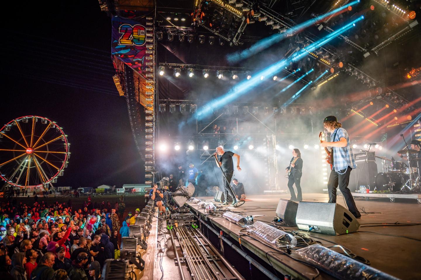 Band performing energetically on stage at night, with festival lights and ferris wheel.