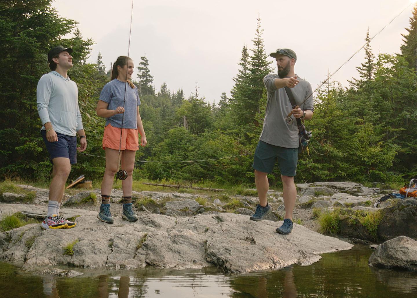 Three people standing on rocks near the water learning how to fish. 