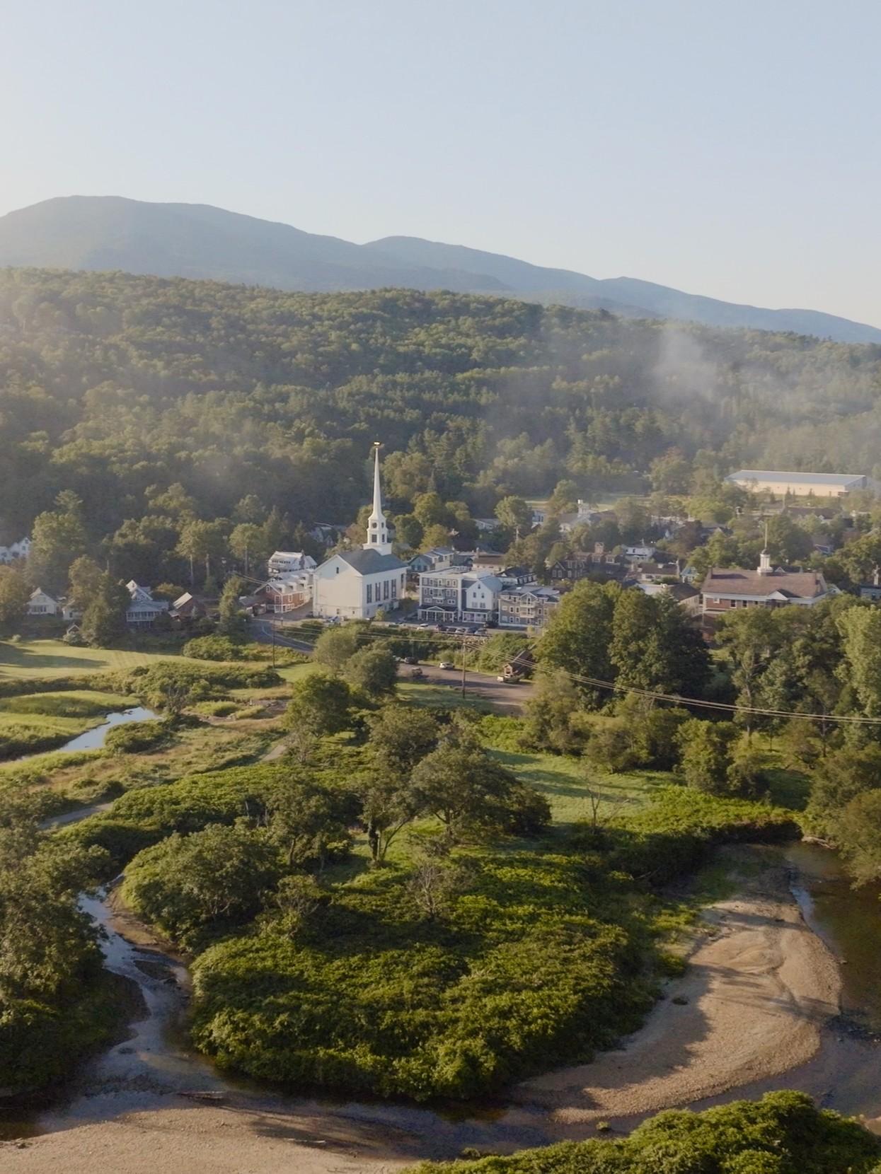 Small village with church spire, surrounded by lush greenery and mountains.