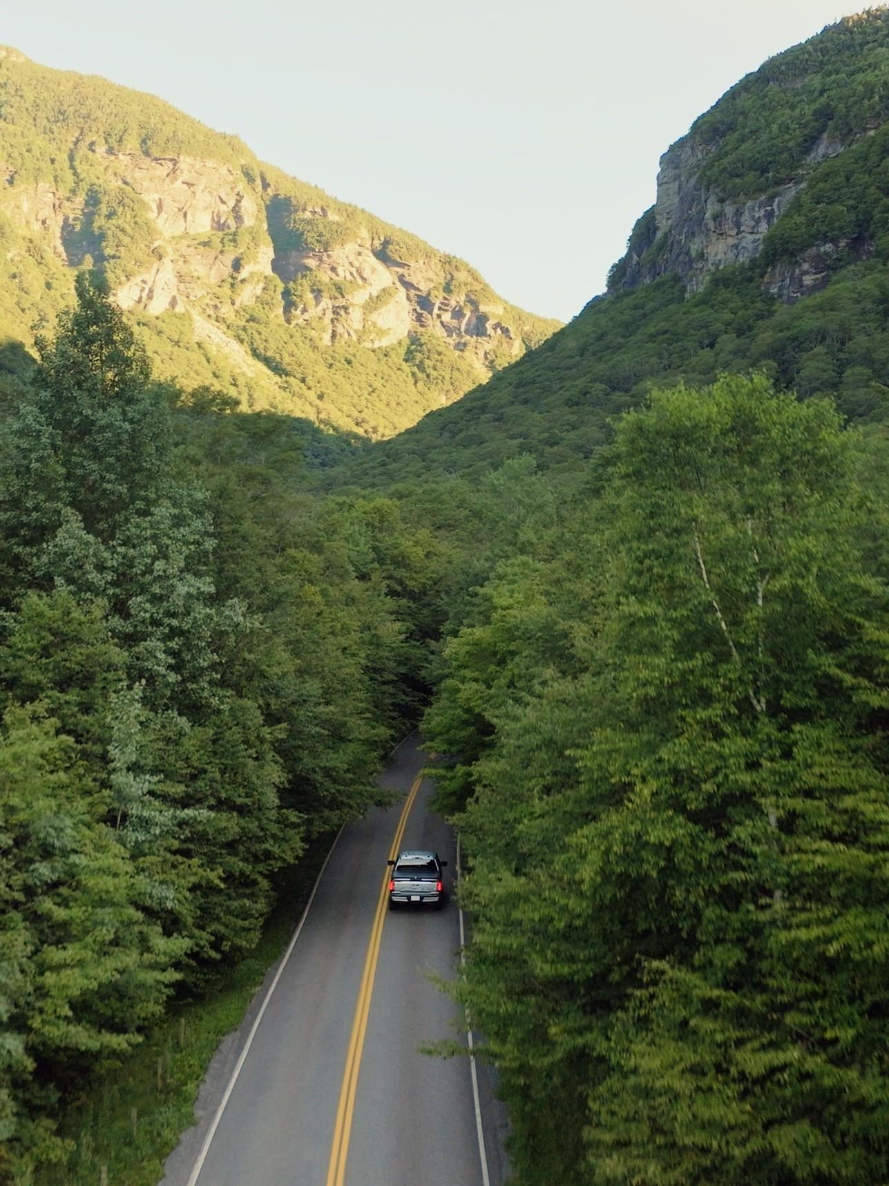 A car drives through a forested mountain pass under a clear sky.