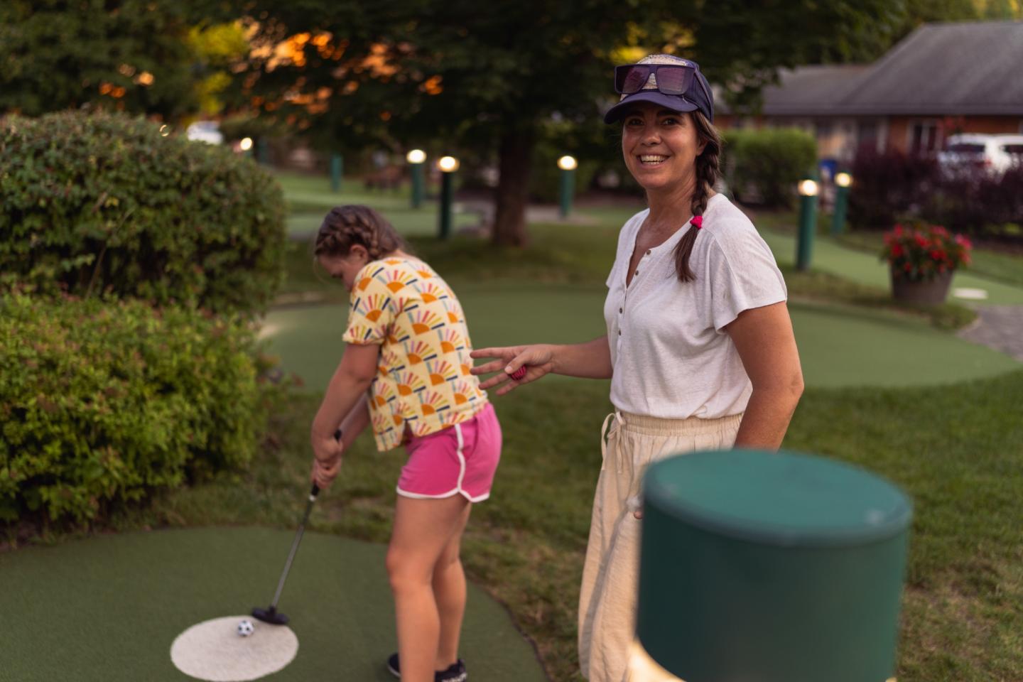 Child playing mini-golf while an adult watches and smiles, outdoors at sunset.
