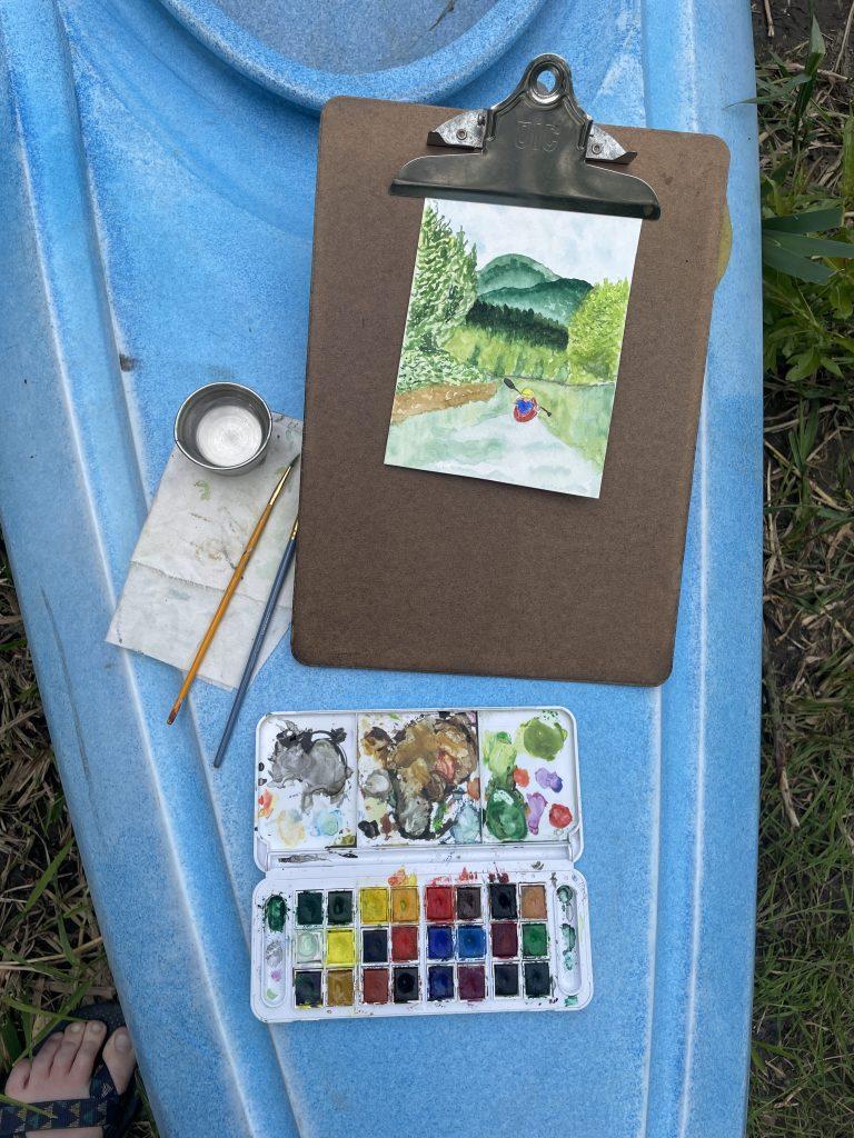 Watercolor painting of landscape on a board with paint palette on blue kayak.
