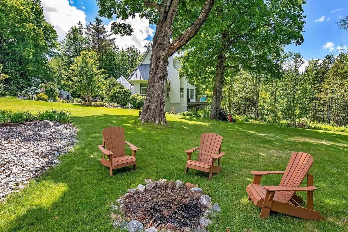 Three Adirondack chairs around a fire pit on a grassy lawn with trees and a house in the background.