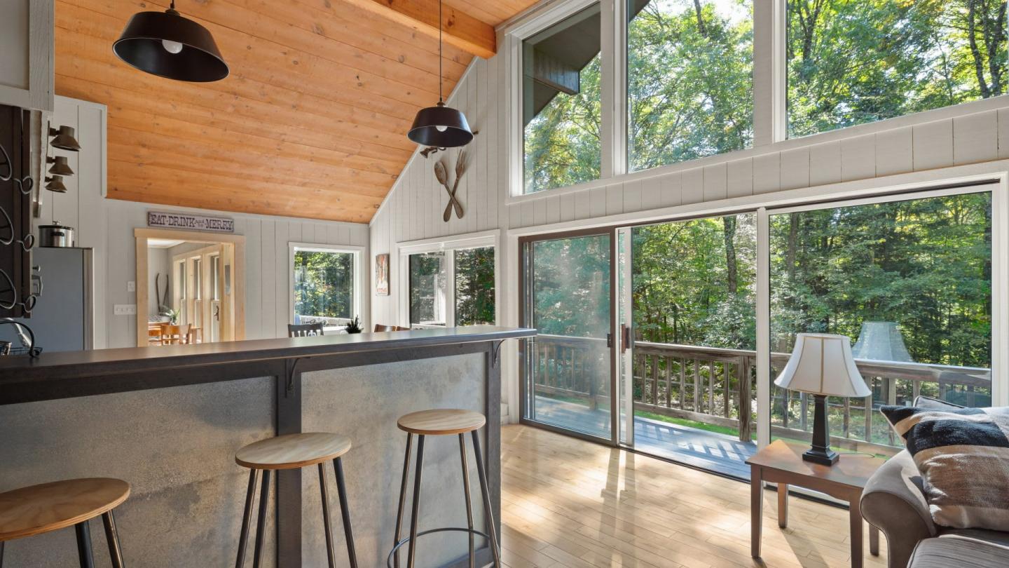 Spacious living room with large windows, wooden ceiling, and bar stools, overlooking trees.