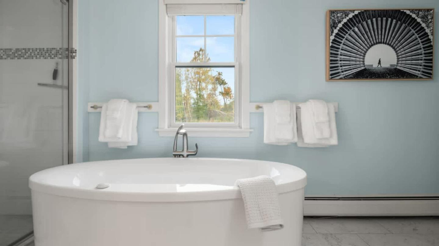 Freestanding white bathtub in a light blue bathroom with a window and modern art.
