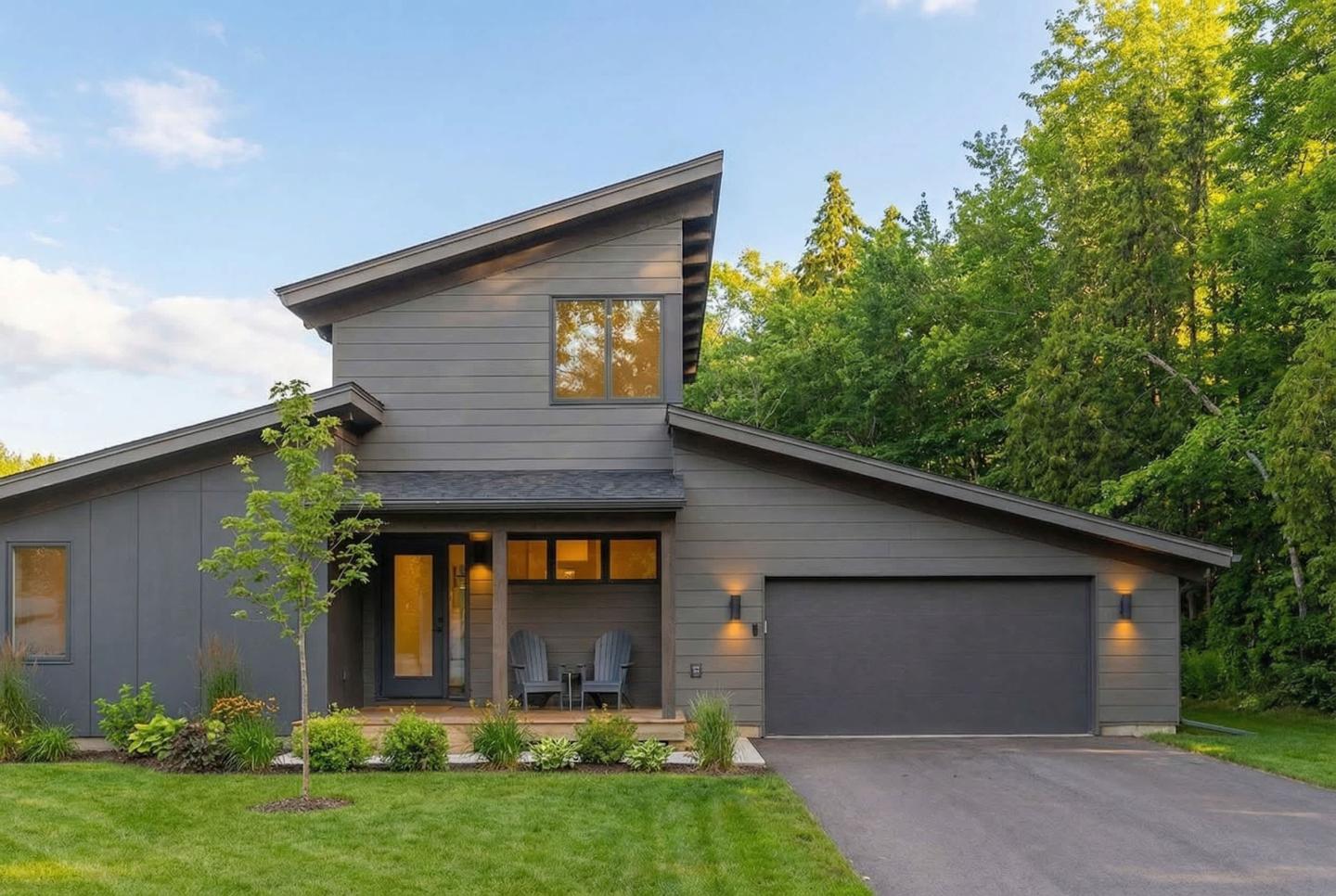 Modern grey house with large windows, surrounded by trees and a lawn, under a blue sky.