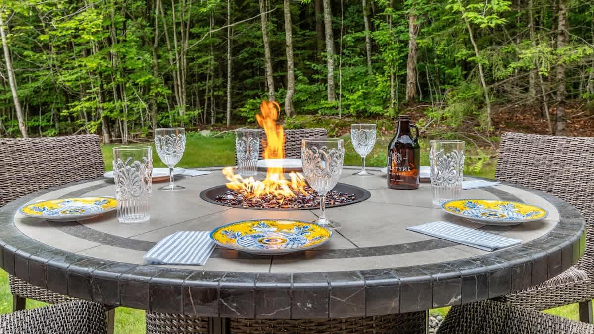Outdoor table with a fire pit, plates, glasses, and a bottle, set in a wooded area.