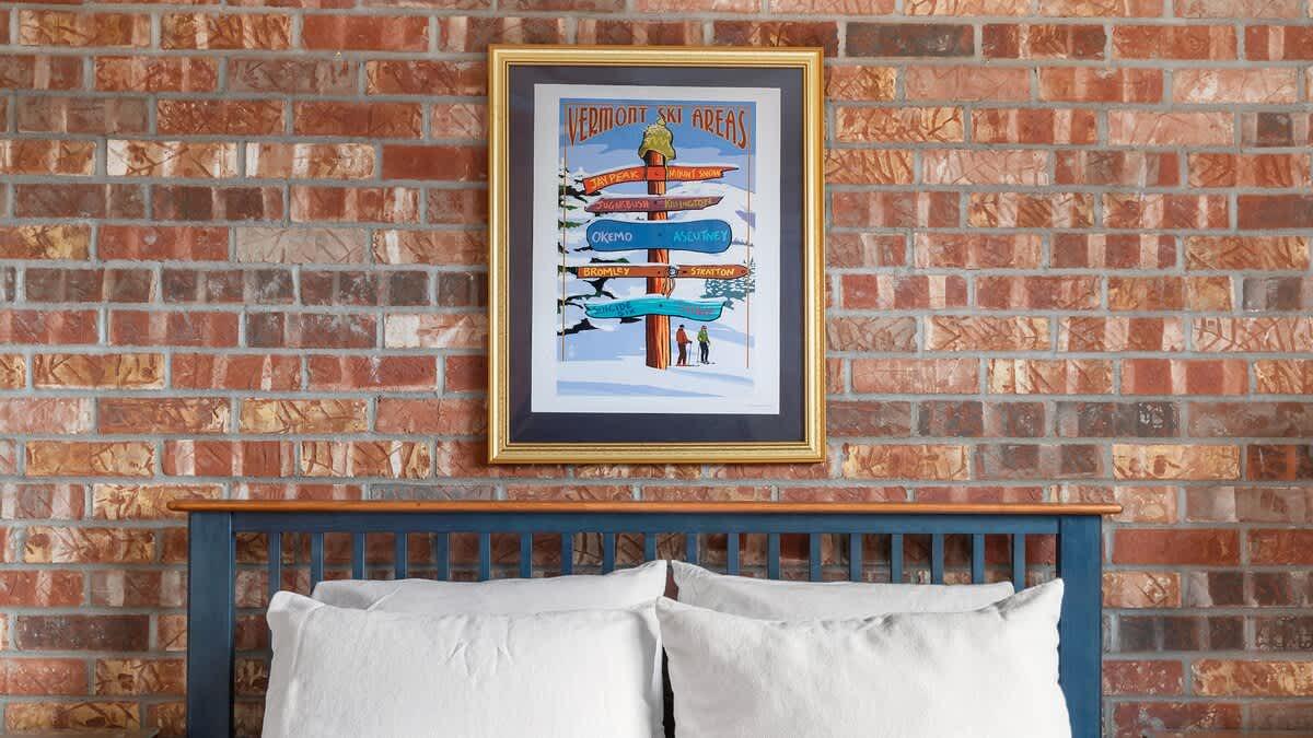Framed poster above a bed against a brick wall.