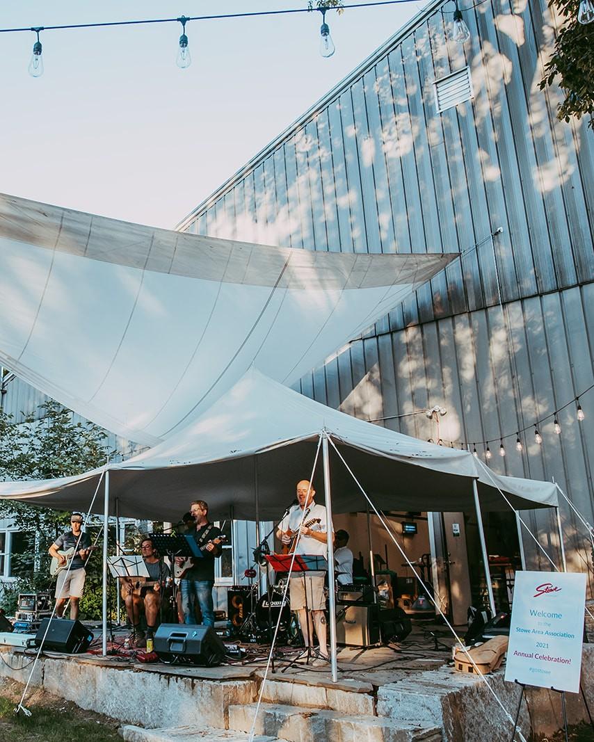 Band performing under a tent on a sunny day with string lights above.