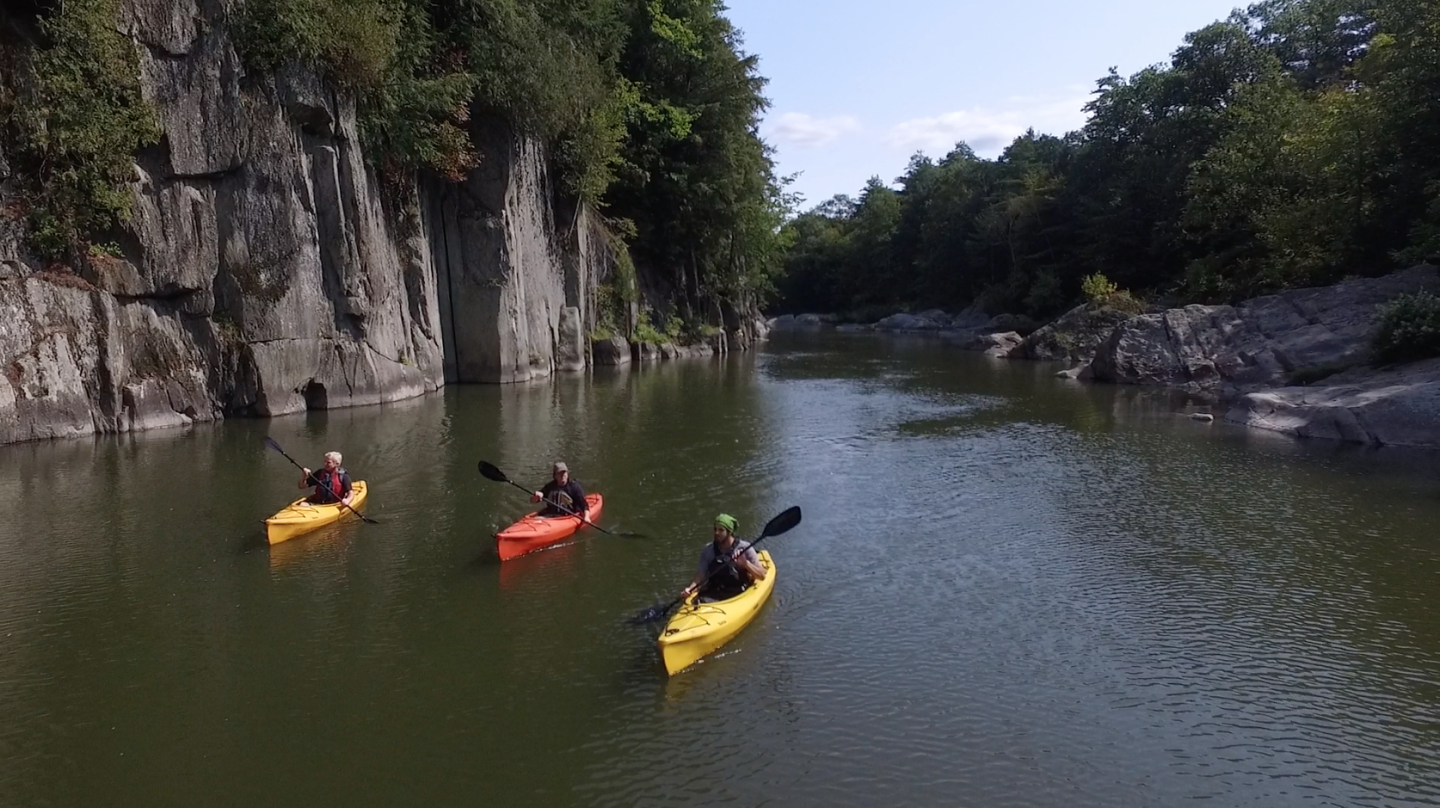 Three kayakers on a calm river with rocky cliffs and trees in the background.